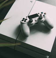 A white gaming console is placed on a dark surface, with a matching white controller resting on top. The console features smooth, clean lines and a sleek design. The surrounding area is slightly blurred, giving focus to the gaming equipment.