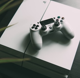 A white gaming console is placed on a dark surface, with a matching white controller resting on top. The console features smooth, clean lines and a sleek design. The surrounding area is slightly blurred, giving focus to the gaming equipment.