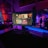 A gaming setup featuring multiple screens with vibrant game-related images illuminated by colorful LED lighting. The environment is dimly lit, highlighting the vivid displays on the monitors. A keyboard is visible on the desk beneath the central monitor.