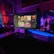 A gaming setup featuring multiple screens with vibrant game-related images illuminated by colorful LED lighting. The environment is dimly lit, highlighting the vivid displays on the monitors. A keyboard is visible on the desk beneath the central monitor.