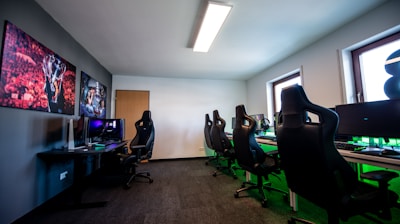 A well-lit gaming room features multiple gaming stations with high-backed chairs and monitors. The room has a minimalist setup with green and black color tones. Large posters adorn the walls, and the room appears clean and organized.
