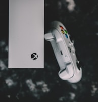 A top view of a white gaming console and a matching controller set against a dark, textured background. The console features a prominent logo, while the controller displays colorful buttons.