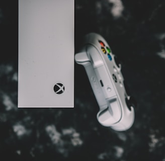 A top view of a white gaming console and a matching controller set against a dark, textured background. The console features a prominent logo, while the controller displays colorful buttons.