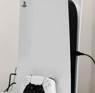 A sleek white gaming console with a modern design, featuring a black logo on its top left. A matching white controller with black accents is placed in front of the console, connected by a cable. The setup is on a plain white surface, creating a minimalist aesthetic.