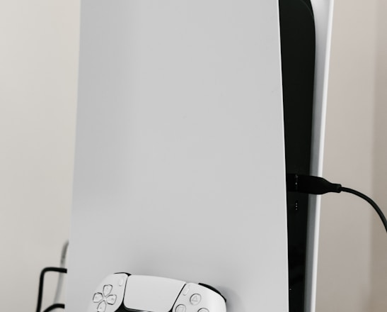 A sleek white gaming console with a modern design, featuring a black logo on its top left. A matching white controller with black accents is placed in front of the console, connected by a cable. The setup is on a plain white surface, creating a minimalist aesthetic.