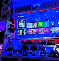 A modern gaming setup featuring a large wall-mounted screen displaying a gaming console interface. The room is decorated with gaming-themed items such as controllers and collectibles. The ambient lighting includes shades of blue and red, creating a vibrant atmosphere.