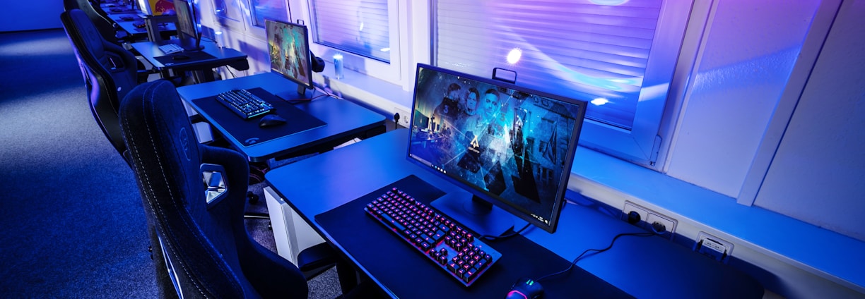 A row of modern gaming setups is arranged neatly along a window-lined wall. Each station includes a high-resolution monitor displaying vivid graphics, a mechanical keyboard with colored lighting, a gaming mouse, and an ergonomic chair. The room is illuminated with ambient blue and purple lights, creating a sleek and futuristic atmosphere.