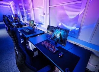 A row of modern gaming setups is arranged neatly along a window-lined wall. Each station includes a high-resolution monitor displaying vivid graphics, a mechanical keyboard with colored lighting, a gaming mouse, and an ergonomic chair. The room is illuminated with ambient blue and purple lights, creating a sleek and futuristic atmosphere.