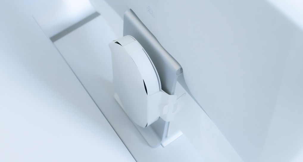 A white, sleek, vertical gaming console placed beside a large, slim monitor. The setting exudes minimalism with a focus on modern technology design.